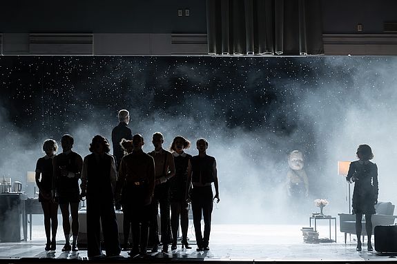 Szenenfoto aus &laquo;Cabaret&raquo;: Im Vordergrund stehen silhouettenhaft die Kit Kat Boys and Girls. Dahinter, von ihnen abgewandt, der Conferencier (Vincent Glander). Rechts steht Sally Bowles (Vassilissa Reznikoff) vor einem Sessel. Die R&uuml;ckwand ist ge&ouml;ffnet, dahinter f&auml;llt ascheartiger &laquo;Schnee&raquo; in einen dunklen Raum.