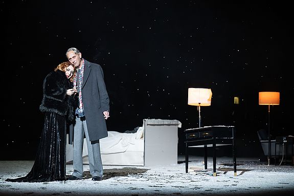 Szenenfoto aus &laquo;Cabaret&raquo;: Sally Bowles (Vassilissa Reznikoff) lehnt sich in schwarzem Kleid und Pelz an Clifford Bradshaw (Michael Goldberg), der neben ihr steht; beide wirken eng verbunden, w&auml;hrend sie gemeinsam in den Raum blicken. Im Hintergrund ein Bett und eine Stehlampe auf der schneebedeckten B&uuml;hne.
