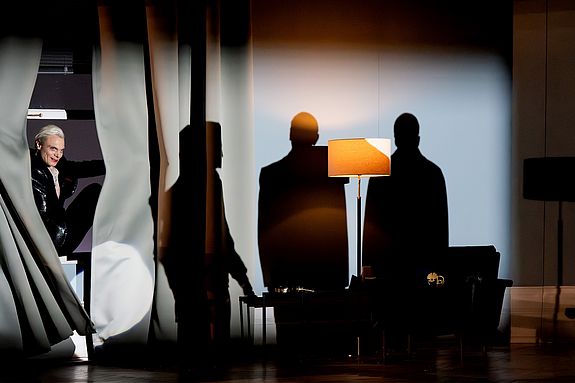 Szenenfoto aus &laquo;Cabaret&raquo;: Der Conferencier (Vincent Glander) erscheint seitlich zwischen Vorh&auml;ngen und blickt ins Publikum. Im Raum dahinter werfen zwei Figuren als Schattenrisse ihre Silhouetten auf die Wand neben einer Stehlampe.