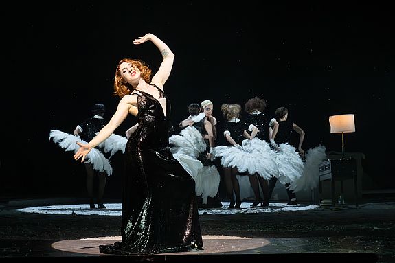 Szenenfoto aus &laquo;Cabaret&raquo;: Sally Bowles (Vassilissa Reznikoff) tanzt im Vordergrund in einem schwarzen Kleid. Hinter ihr stehen die Kit Kat Girls und Boys in Kost&uuml;men mit Federn; unter ihnen der Conferencier (Vincent Glander), der zu ihr blickt.