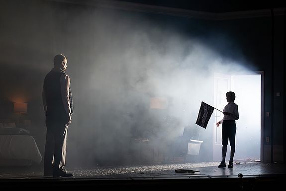 Szenenfoto aus &laquo;Cabaret&raquo;: Clifford Bradshaw (Michael Goldberg) steht in einem abgedunkelten Raum einem Jungen gegen&uuml;ber, der in einer hell erleuchteten T&uuml;r&ouml;ffnung steht und eine Fahne h&auml;lt; Nebel erf&uuml;llt die B&uuml;hne.