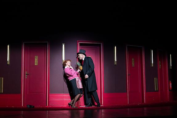 Szenenfoto aus &laquo;Cabaret&raquo;: Fr&auml;ulein Schneider (Cathrin St&ouml;rmer) und Herr Schultz (Robert D&ouml;lle) stehen sich auf einem Flur mit mehreren roten T&uuml;ren gegen&uuml;ber. Er h&auml;lt eine Ananas in der Hand, w&auml;hrend sie sich l&auml;chelnd an die Wand lehnt.