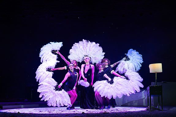 Szenenfoto aus &laquo;Cabaret&raquo;: Der Conf&eacute;rencier steht mit T&auml;nzer*innen des Kit Kat Klub auf der B&uuml;hne, umgeben von gro&szlig;en wei&szlig;en Federf&auml;chern vor dunklem Hintergrund.