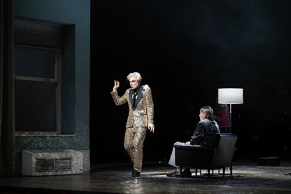 Szenenfoto aus &laquo;Cabaret&raquo;: Der Conf&eacute;rencier (Vincent Glander) in glitzerndem Anzug bewegt sich &uuml;ber die B&uuml;hne; im Hintergrund sitzt Clifford Bradshaw (Michael Goldberg) in einem Sessel, von uns abgewandt, w&auml;hrend wei&szlig;e Partikel wie Schnee herabfallen.